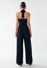 Tamsiai mėlynas kombinezonas su pakabinamomis petnešomis, prigludusiu viršumi, plačiomis kelnėmis, užtrauktuku nugarinėje dalyje ir kaklo juostele, pasižymintis lygiu, blizgančiu paviršiumi.