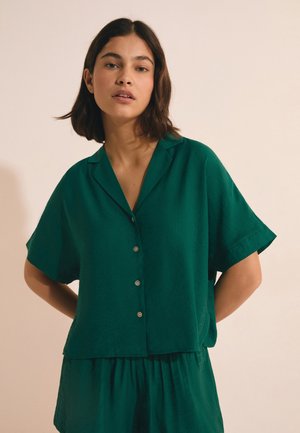 Joven mujer con cabello oscuro hasta los hombros, vestida con una camisa verde de manga corta con botones y falda a juego, de pie con las manos detrás de la espalda.