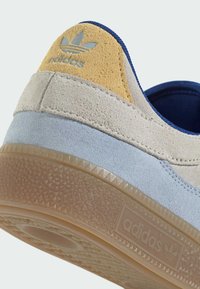 Adidas sneaker featuring a beige, blue, and yellow suede upper with textured gum sole. Accented with an embossed logo and stitching details.