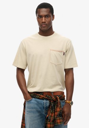 Beige katoenen t-shirt met een borstzakje met rode stitching. Gerdragen met blauwe jeans en een geruite shirt om de taille geknoopt.