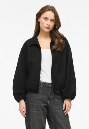Kvinde med langt brunt hår iført en sort lynlåsjakke over en hvid top og mørkegrå jeans, stående mod en hvid baggrund.