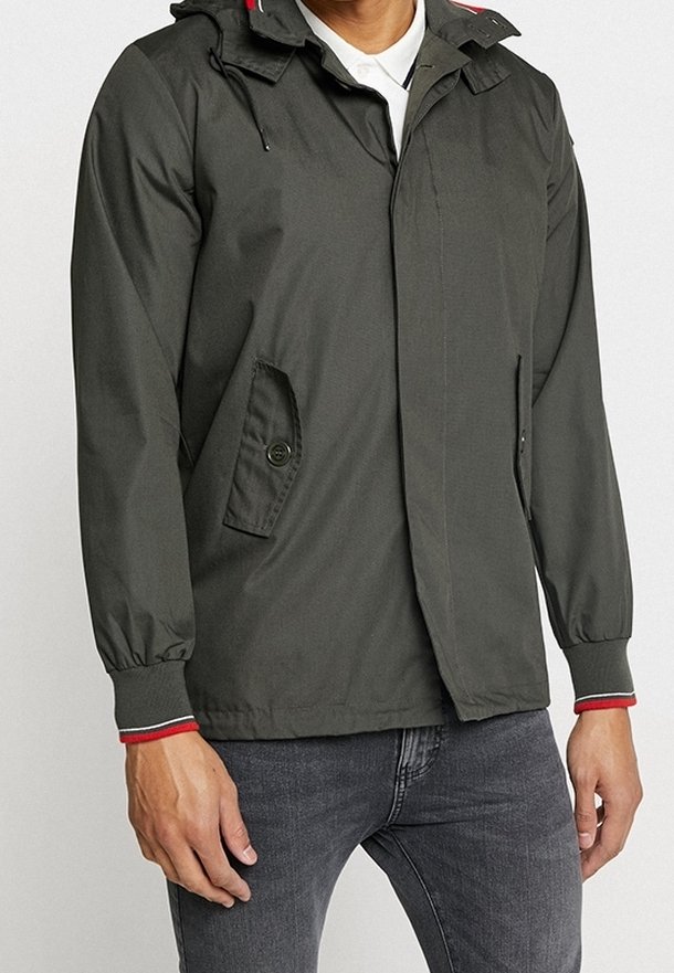 Donkergroene waterdichte jas met capuchon, ritssluiting aan de voorkant, zijzakken, elastische manchetten en rode accenten op de capuchon en manchetten.