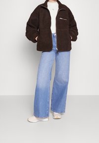 Brązowa polarowa kurtka z zamykaną kieszenią, zestawiona z jasnoniebieskimi spodniami dzwonami. Kremowe sneakersy dopełniają wygląd. Widoczna miękka tekstura.