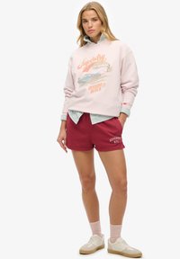 Lichtroze sweatshirt met 'Superdry' opdruk, gecombineerd met bordeauxrode shorts met logo. Het model draagt witte sneakers en roze sokken.
