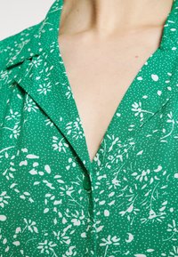 Grünes Hemd mit floralem Druck, das weiße Muster, einen V-Ausschnitt und eine Knopfleiste aufweist. Der Stoff hat ein strukturiertes Erscheinungsbild mit einem punktierten Hintergrund.