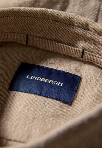 Bruine stof met een gestructureerde afwerking, met een marineblauw label dat "LINDBERGH" in witte letters leest, genaaid in de hals van het kledingstuk.