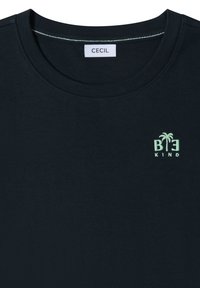 T-shirt noir à col rond avec texte vert "BE KIND" et motif de palmier sur le côté gauche de la poitrine, étiqueté "CECIL" sur le col intérieur.