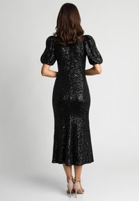 Czarna sukienka z cekinami o dopasowanym kroju i bufiastych rękawach, zwężająca się ku rozszerzanemu dołowi. Noszona z jasnymi butami na wysokim obcasie.