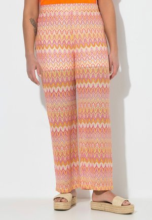 Frau trägt hoch taillierte, weit geschnittene Hosen mit Zickzackmustern in Pink, Orange und Gelb sowie beigefarbene Plateausandalen und steht vor einer weißen Wand.