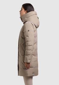 Béžový zateplený zimní kabát s vysokým límcem, velkou kapucí, bočními kapsami a elegantním designem. Obsahuje brandingovou nášivku na rukávu.