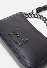 Pochette en faux cuir noir avec surface texturée, quincaillerie métallique et logo en relief. Dispose d'une bandoulière chaîne et d'une forme rectangulaire.