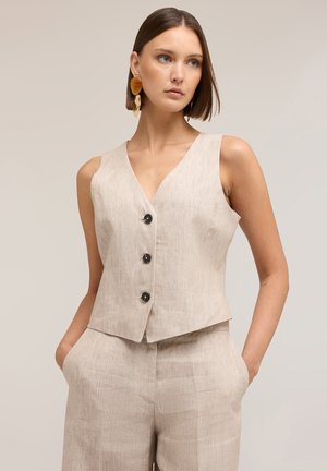 Frau trägt beige ärmellose, knöpfbare Weste und passende hochgeschnittene Hose, mit kurzem glattem Haar und großen bernsteinfarbenen Ohrringen, Hände in den Taschen.