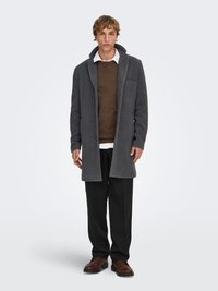 Manteau en laine grise avec col cranté, une poche poitrine, porté sur un pull marron et une chemise blanche. Pantalon noir et chaussures marron complètent la tenue.