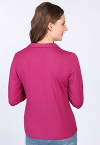 Fuchsia Langarmhemd mit Kragen, glatter Textur und einer geschwungenen Nahtdetails an der Rückseite. Kombiniert mit hellblauen Jeans.