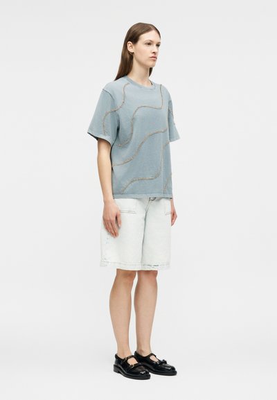 Γυναίκα που στέκεται φορώντας μπλε πουκάμισο με σχέδια και κοντά μανίκια, ανοιχτόχρωμα τζιν σορτς μέχρι το γόνατο και μαύρα παπούτσια τύπου Mary Jane.