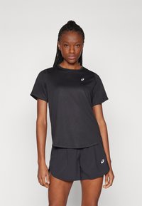 Donna con capelli intrecciati che indossa una maglietta atletica nera a maniche corte e pantaloncini con loghi bianchi, in piedi contro uno sfondo chiaro e uniforme.