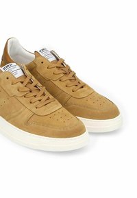 Baskets en suede beige avec des détails perforés et des lacets plats. Semelles en caoutchouc blanc avec une finition texturée. Étiquette sur la languette affichant le nom de la marque.