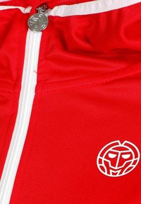 Chaqueta roja con cremallera blanca y un logo blanco que asemeja un rostro estilizado en el pecho, con un tirador de cremallera circular plateado.
