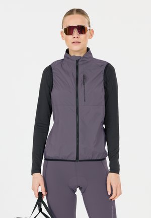 Frau trägt lila Fahrradweste und Leggings, schwarze langärmelige Basisschicht, große reflektierende Sportsonnenbrille, hält Helmriemen fest.