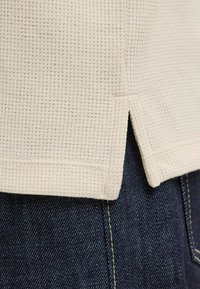 Gros plan sur l'ourlet supérieur en tissu gaufré beige avec fente latérale sur un jean en denim bleu foncé avec coutures visibles.