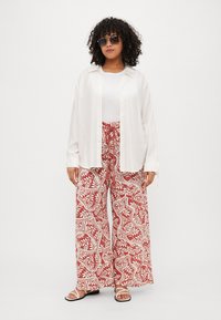 Cămașă albă cu mâneci lungi peste un top alb fără mâneci, asociată cu pantaloni largi roșii și albi cu un model și sandale plate.