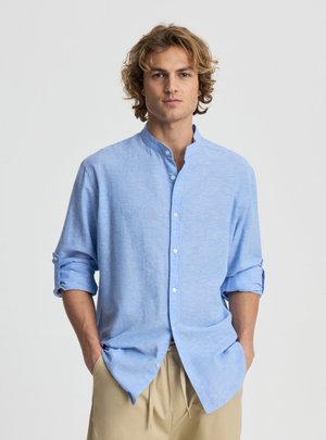 Młody mężczyzna z blond kręconymi włosami stoi z rękami w kieszeniach, ubrany w jasnoniebieską koszulę zapinaną na guziki z podwiniętymi rękawami i beżowe spodnie.