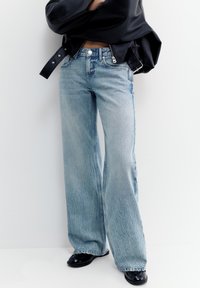 Személy világoskék bő szárú farmernadrágban, fekete bőrdzsekiben és fekete cipőben, fehér falnak támaszkodva.