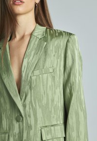 Blazer verde in raso con un sottile motivo a onde, rever a punta, chiusura con un solo bottone e due tasche frontali. Texture liscia.