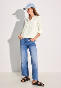 Lys olivengrøn tekstureret pullover med henley-udskæring, blå denim brede bukser og sorte sandaler med guldspænder, kompletteret med en lyseblå kasket.