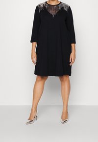 Svart klänning med trekvarts ärmar, med en halsringning prydd med paljetter. Tyget är mjukt och formen är löst sittande. Silverfärgade klackskor.