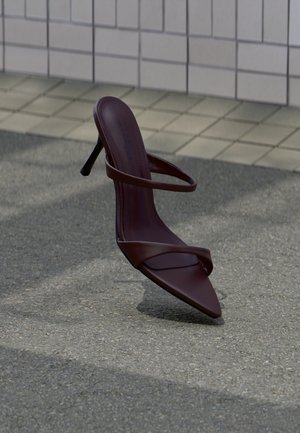 Sandale à talon haut brun foncé avec des lanières fines, flottant au-dessus d'un bitume gris près d'un mur carrelé.