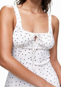 Vestido blanco con lunares negros, escote cuadrado y tela fruncida. Presenta un detalle de lazo ajustable en el frente y tirantes anchos.