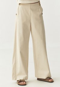 Pantalons larges beiges en coton, dotés de grands boutons sur le côté et d'une texture douce. Coupés à la cheville avec une coupe décontractée.