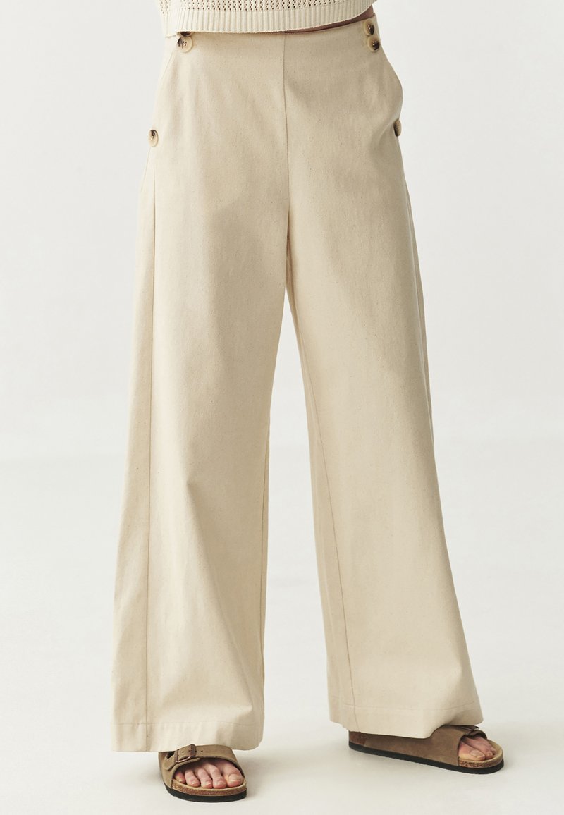 Pantalons larges beiges en coton, dotés de grands boutons sur le côté et d'une texture douce. Coupés à la cheville avec une coupe décontractée.