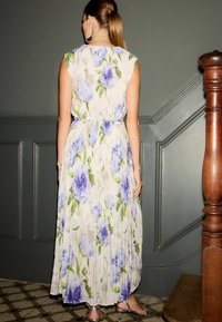 Áramló, pliszírozott, világos bézs maxiruha kék virágmintával, ujjatlan kivitel, összeszorított derékkal és rétegelt szegéllyel. Ezüst, pántos cipők.