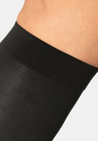 Schwarze Beinwärmer aus strukturiertem Material mit einem glatten, kontrastierenden Bündchen oben für einen sicheren Sitz. Nahaufnahme zeigt die Details.