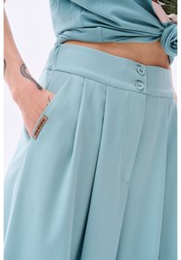 Jupe plissée bleu clair en tissu doux, avec deux boutons à la taille et une poche latérale arborant une étiquette de marque.