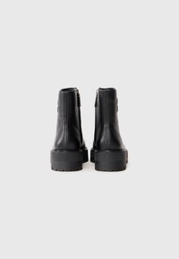 Une paire de bottes noires en cuir chunky, mi-mollet, avec des semelles épaisses et rainurées, et des zips à l'arrière, présentées de dos sur un fond blanc.