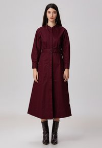 Robe chemise longue bordeaux avec une ceinture, manches longues, fermeture à boutons et coupe droite. Portée avec des bottines noires brillantes.
