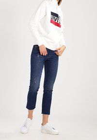 Sweat à capuche blanc avec un logo Levi's rouge et noir, associé à un jean bleu foncé déchiré et des baskets blanches. Tenue décontractée, avec capuche et poches.