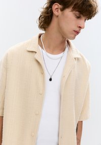 Beige košeľa s krátkym rukávom a gombíkmi, s textúrovanými pruhmi, nosená cez biely priliehavý top. Doplnková vrstva náhrdelníkov.