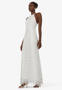 Witte kanten maxi-jurk met een halterhals, bloemenpatroon en golvende zoom. G gedragen met strappy witte hakken en heeft een aansluitende top.