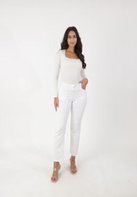 Top de manga larga y escote cuadrado en beige claro, combinado con jeans blancos de pierna recta y sandalias de tacón transparente, sobre un fondo liso.