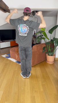 Šedé tričko s barevnou grafikou hory na zadní straně, v kombinaci se šedými džíny a bílými teniskami. Vnitřní prostředí s rostlinami.