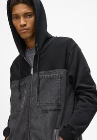 Μαύρη κουκούλα με αντίθετο γκρι πάνελ από ντένιμ, κλείσιμο με φερμουάρ, δύο μπροστινές τσέπες, ρυθμιζόμενα κορδόνια και λεπτομέρειες λογότυπου στο στήθος.