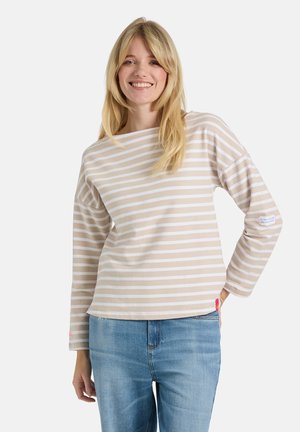Young woman smiling, wearing beige and white striped long-sleeve top and blue jeans, with one hand resting on her hip.