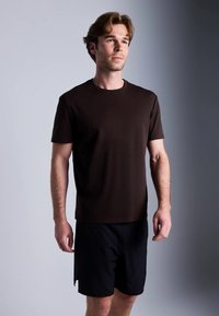 Ung mann med lys brunt hår iført en enkel mørkebrun T-skjorte og svarte shorts, som står mot en gradient grå bakgrunn.