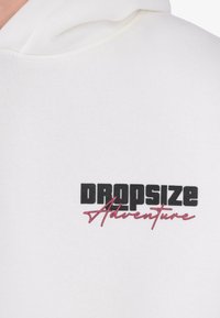 Hvid hættejakke med fed sort tekst "Dropsize" og lyserød kursiv tekst "Adventure" trykt på brystområdet.