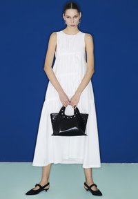 Vestido blanco sin mangas con cintura fruncida y falda fluida, combinado con un bolso negro con tachuelas y asas, sobre un fondo azul.