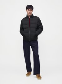 Homme debout, les mains dans les poches, portant une doudoune noire, un pantalon foncé, des baskets marron et une chemise rouge sur un fond blanc uni.